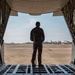 A military service member in camouflage uniform stands at the open rear cargo ramp of a large military transport aircraft, looking out onto an airfield tarmac. The perspective is from inside the aircraft looking outward, creating a dramatic framing effect with the cargo bay walls and ramp. Additional military aircraft and structures are visible in the background on the flight line.