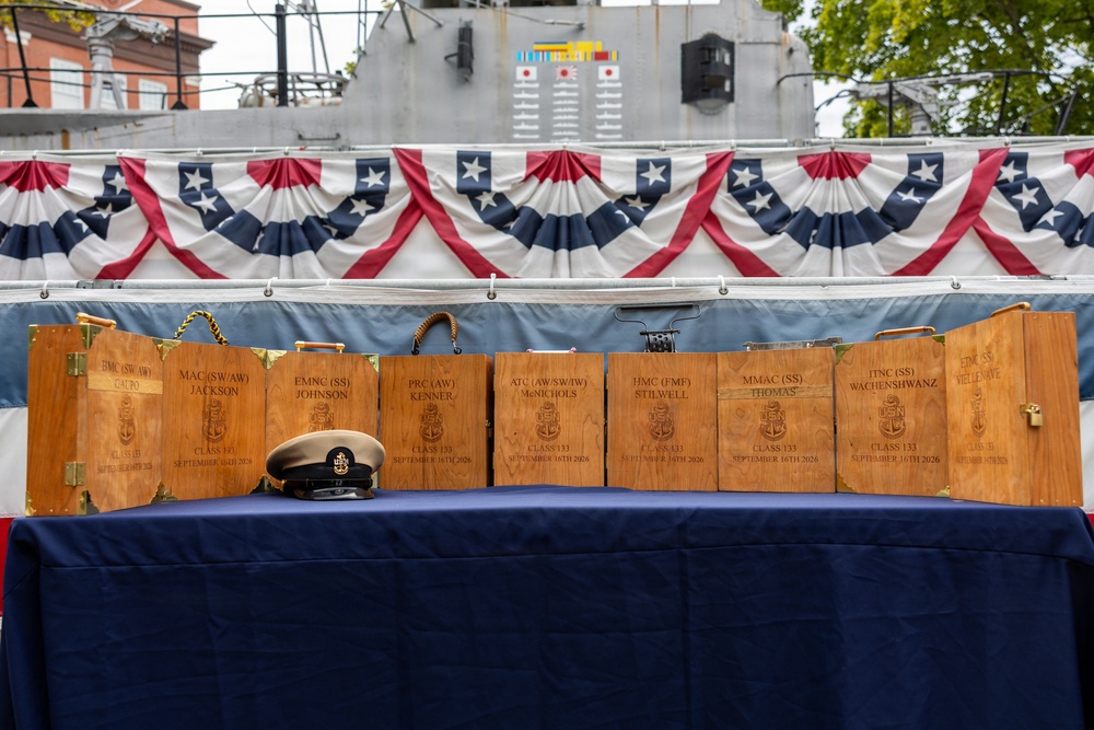 A ceremonial display table covered in a blue cloth holds multiple engraved wooden boxes/cases arranged in a row, with a Navy chief petty officer's combination cover (hat) placed in front of them. The boxes appear to be personalized chief petty officer initiation or advancement ceremony cases, each bearing individual names, ratings, and a date of September 16th, 2026, Class 133. The setting is decorated with patriotic red, white, and blue bunting, with a naval vessel visible in the background.