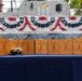 A ceremonial display table covered in a blue cloth holds multiple engraved wooden boxes/cases arranged in a row, with a Navy chief petty officer's combination cover (hat) placed in front of them. The boxes appear to be personalized chief petty officer initiation or advancement ceremony cases, each bearing individual names, ratings, and a date of September 16th, 2026, Class 133. The setting is decorated with patriotic red, white, and blue bunting, with a naval vessel visible in the background.
