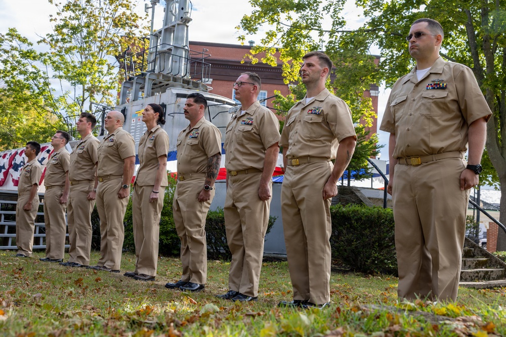 A group of U.S. Navy personnel in tan/khaki service uniforms are standing at attention in a line during what appears to be an outdoor military ceremony. They are positioned on a grass area in front of what appears to be a naval vessel or boat display, with a brick building and trees visible in the background. Several of the personnel display ribbons and rank insignia on their uniforms.