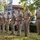 A group of U.S. Navy personnel in tan/khaki service uniforms are standing at attention in a line during what appears to be an outdoor military ceremony. They are positioned on a grass area in front of what appears to be a naval vessel or boat display, with a brick building and trees visible in the background. Several of the personnel display ribbons and rank insignia on their uniforms.