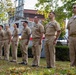 A group of U.S. Navy personnel in tan/khaki service uniforms are standing at attention in a line during what appears to be an outdoor military ceremony. They are positioned on a grass area in front of what appears to be a naval vessel or boat display, with a brick building and trees visible in the background. Several of the personnel display ribbons and rank insignia on their uniforms.