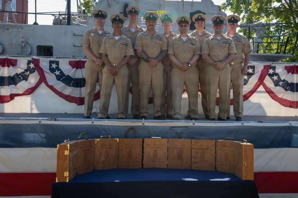 A group of approximately nine military personnel, predominantly in U.S. Navy khaki service uniforms, pose for a formal group photograph in front of a naval vessel decorated with patriotic bunting. In the foreground, several wooden presentation boxes are displayed on a table draped with a dark blue cloth. The setting suggests a formal military ceremony, likely a promotion, retirement, or award event.