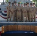 A group of approximately nine military personnel, predominantly in U.S. Navy khaki service uniforms, pose for a formal group photograph in front of a naval vessel decorated with patriotic bunting. In the foreground, several wooden presentation boxes are displayed on a table draped with a dark blue cloth. The setting suggests a formal military ceremony, likely a promotion, retirement, or award event.