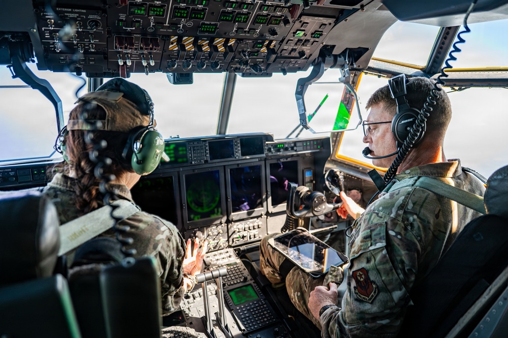 Two military aircrew members are seated in the cockpit of a large military transport aircraft, likely a C-130 or similar platform, during an active flight. Both personnel are wearing military camouflage uniforms and aviation headsets, with one holding a tablet device. The cockpit displays numerous digital avionics screens, overhead panels, and flight instruments consistent with a modern military transport aircraft.
