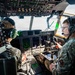 Two military aircrew members are seated in the cockpit of a large military transport aircraft, likely a C-130 or similar platform, during an active flight. Both personnel are wearing military camouflage uniforms and aviation headsets, with one holding a tablet device. The cockpit displays numerous digital avionics screens, overhead panels, and flight instruments consistent with a modern military transport aircraft.