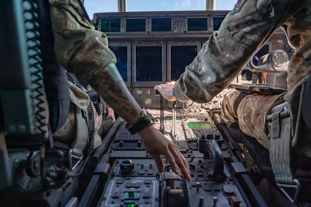 Two military aircrew members in camouflage uniforms are operating controls in a large aircraft cockpit, likely a military transport or tanker aircraft. The image is taken from a low center-console perspective between the two pilots, showing the instrument panel, throttle levers, and multiple digital displays. One crew member has a visible tattoo on their forearm.