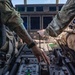 Two military aircrew members in camouflage uniforms are operating controls in a large aircraft cockpit, likely a military transport or tanker aircraft. The image is taken from a low center-console perspective between the two pilots, showing the instrument panel, throttle levers, and multiple digital displays. One crew member has a visible tattoo on their forearm.