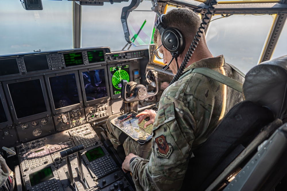 A military aviator in camouflage uniform is seated in the cockpit of a large military aircraft, reviewing what appears to be a navigational chart or map on a tablet/kneeboard. The cockpit features an advanced glass cockpit configuration with multiple digital displays, green-lit avionics screens, and a radar display, consistent with a C-130J Super Hercules or similar large military transport aircraft. The aircraft is airborne, as evidenced by sky and clouds visible through the cockpit windows.