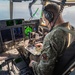 A military aviator in camouflage uniform is seated in the cockpit of a large military aircraft, reviewing what appears to be a navigational chart or map on a tablet/kneeboard. The cockpit features an advanced glass cockpit configuration with multiple digital displays, green-lit avionics screens, and a radar display, consistent with a C-130J Super Hercules or similar large military transport aircraft. The aircraft is airborne, as evidenced by sky and clouds visible through the cockpit windows.