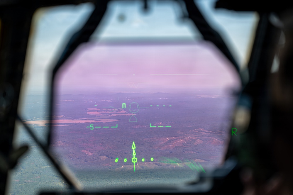 The image shows a view through a military aircraft's Head-Up Display (HUD) with green symbology overlaid on a landscape below. The HUD glass has a distinctive pink/purple anti-reflective coating tint, and the green phosphor display shows flight instrumentation symbols. The terrain visible below consists of forested hills, open fields, and what appear to be bodies of water.