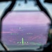 The image shows a view through a military aircraft's Head-Up Display (HUD) with green symbology overlaid on a landscape below. The HUD glass has a distinctive pink/purple anti-reflective coating tint, and the green phosphor display shows flight instrumentation symbols. The terrain visible below consists of forested hills, open fields, and what appear to be bodies of water.