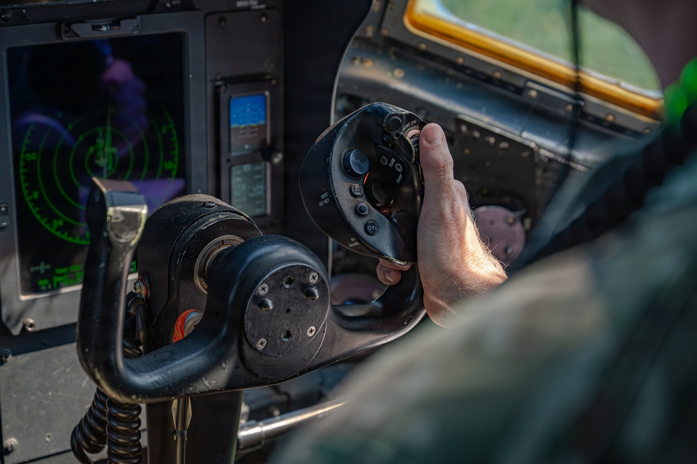 A close-up photograph of a military helicopter cockpit showing a pilot's hand gripping a cyclic control stick with multiple buttons and switches on the grip. The background reveals avionics displays including a green radar/navigation screen and what appears to be a glass cockpit display. The image captures an active flight or pre-flight scenario with detailed cockpit instrumentation visible.