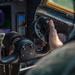 A close-up photograph of a military helicopter cockpit showing a pilot's hand gripping a cyclic control stick with multiple buttons and switches on the grip. The background reveals avionics displays including a green radar/navigation screen and what appears to be a glass cockpit display. The image captures an active flight or pre-flight scenario with detailed cockpit instrumentation visible.