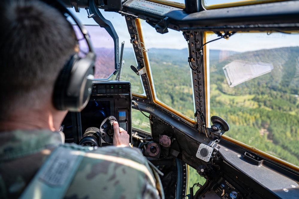 A military pilot in camouflage uniform and aviation headset is operating an aircraft in flight over a forested, mountainous landscape. The cockpit features vintage or older-generation instrumentation with yellow-framed windows, suggesting a legacy military transport or utility aircraft. The terrain below shows green valleys and tree-covered hills visible through the large cockpit windscreen.