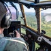 A military pilot in camouflage uniform and aviation headset is operating an aircraft in flight over a forested, mountainous landscape. The cockpit features vintage or older-generation instrumentation with yellow-framed windows, suggesting a legacy military transport or utility aircraft. The terrain below shows green valleys and tree-covered hills visible through the large cockpit windscreen.