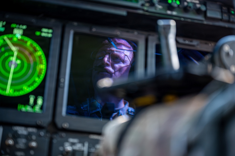 A crew member or pilot is visible in the cockpit of a military or large aircraft, reflected or seen through instrumentation panels. The scene is illuminated primarily by the green glow of a radar display and blue-tinted cockpit lighting. The individual is wearing glasses and appears to be monitoring flight instruments.