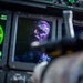 A crew member or pilot is visible in the cockpit of a military or large aircraft, reflected or seen through instrumentation panels. The scene is illuminated primarily by the green glow of a radar display and blue-tinted cockpit lighting. The individual is wearing glasses and appears to be monitoring flight instruments.