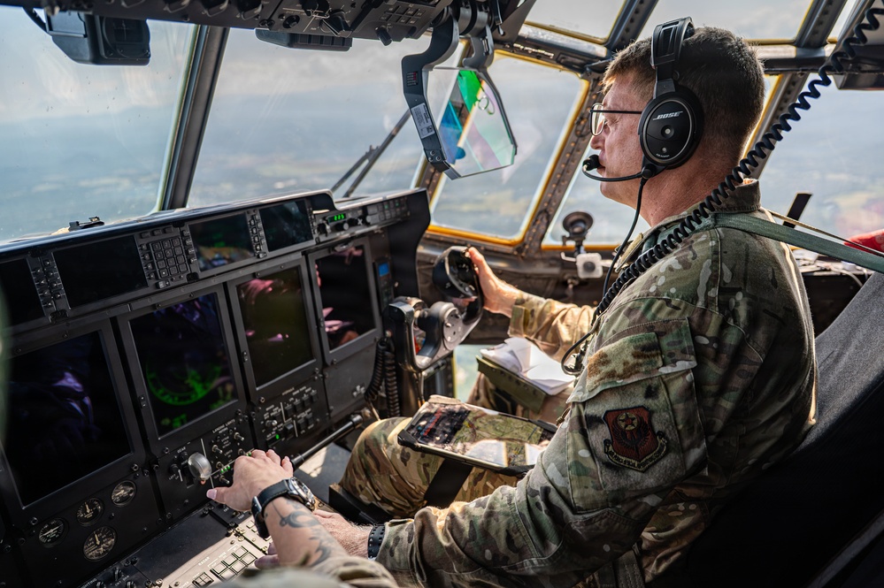 A U.S. Air Force airman is seated in the cockpit of a military transport aircraft, actively operating flight controls at altitude. The pilot is wearing OCP (Operational Camouflage Pattern) uniform with a unit patch visible on the sleeve, Bose aviation headset, and glasses. Multiple digital flight displays and instrument panels are visible in the modern glass cockpit.