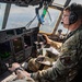 A U.S. Air Force airman is seated in the cockpit of a military transport aircraft, actively operating flight controls at altitude. The pilot is wearing OCP (Operational Camouflage Pattern) uniform with a unit patch visible on the sleeve, Bose aviation headset, and glasses. Multiple digital flight displays and instrument panels are visible in the modern glass cockpit.
