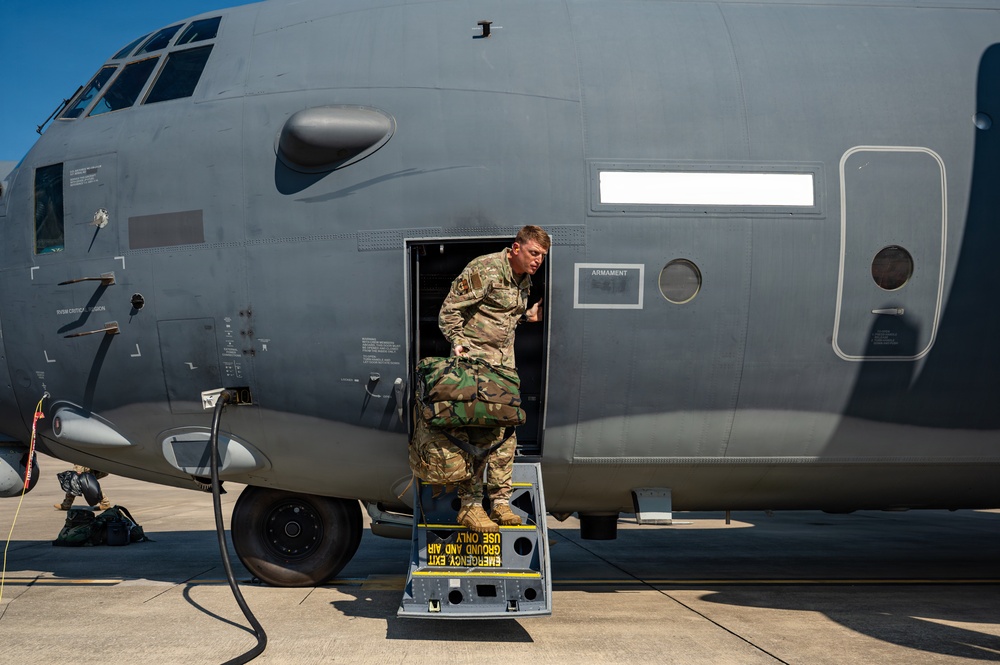 A military service member in MultiCam/OCP camouflage uniform is exiting a large gray military transport aircraft through a side door, carrying a camouflage-patterned bag or equipment. The aircraft appears to be a C-130 Hercules or similar large military transport, with a visible 'ARMAMENT' placard and an 'EMERGENCY EXIT - GROUND AND AIR USE ONLY' warning on the yellow-striped steps. The scene takes place on a tarmac under clear blue skies.