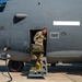 A military service member in MultiCam/OCP camouflage uniform is exiting a large gray military transport aircraft through a side door, carrying a camouflage-patterned bag or equipment. The aircraft appears to be a C-130 Hercules or similar large military transport, with a visible 'ARMAMENT' placard and an 'EMERGENCY EXIT - GROUND AND AIR USE ONLY' warning on the yellow-striped steps. The scene takes place on a tarmac under clear blue skies.
