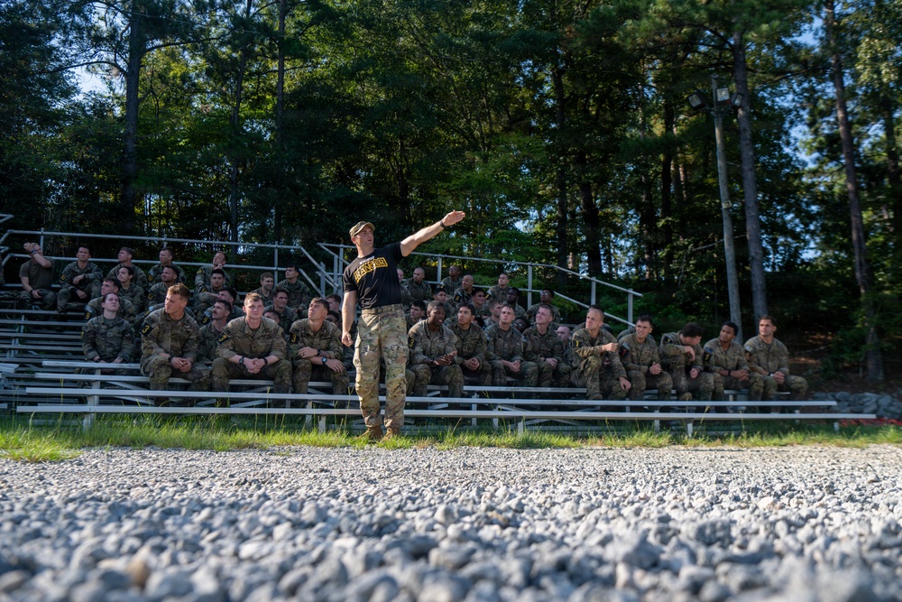 A military instructor or cadre member in a black t-shirt and camouflage pants stands on a gravel area addressing a large group of soldiers seated in bleachers. The soldiers are wearing camouflage uniforms and appear to be attentively listening to a briefing or instruction. The setting appears to be an outdoor training area surrounded by trees.