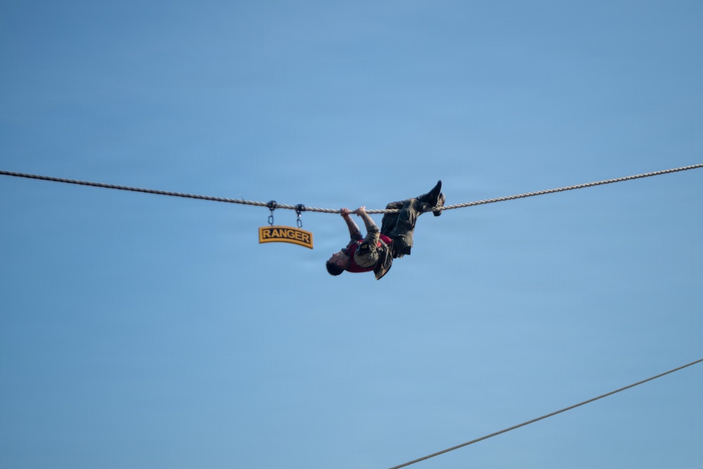 A person is traversing a rope hand-over-hand while hanging upside down against a clear blue sky. A yellow and black 'RANGER' tab sign is attached to the rope nearby, suggesting this is a U.S. Army Ranger training or demonstration event. The individual is wearing military-style clothing and appears to be performing a rope crossing technique.