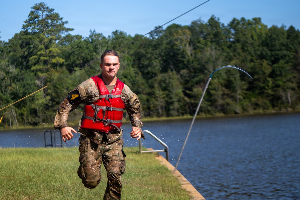 A soldier in camouflage uniform and red life vest runs along the bank of a lake, visibly wet from recent water immersion. The setting appears to be a military training exercise involving water obstacles, with a zip line cable visible overhead and a metal railing leading into the water. The soldier displays a determined expression and is marked with the number '4', suggesting a competitive or evaluated event.