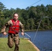 A soldier in camouflage uniform and red life vest runs along the bank of a lake, visibly wet from recent water immersion. The setting appears to be a military training exercise involving water obstacles, with a zip line cable visible overhead and a metal railing leading into the water. The soldier displays a determined expression and is marked with the number '4', suggesting a competitive or evaluated event.