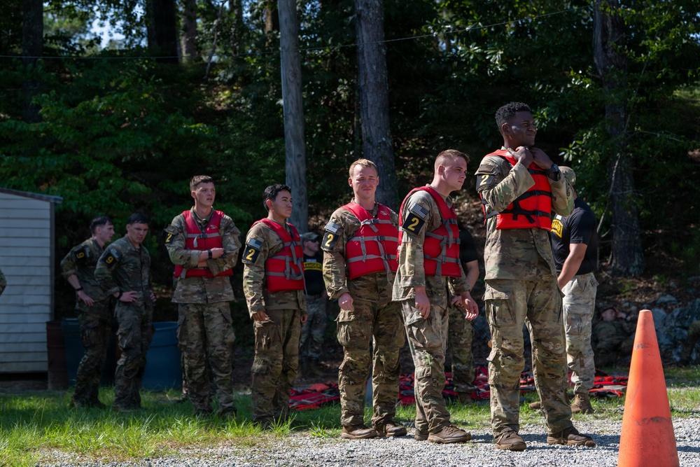 A group of military personnel in camouflage uniforms are standing in a line outdoors, wearing red life vests/personal flotation devices marked with the number '2'. They appear to be participating in a water training exercise or safety briefing near a wooded area. An instructor or cadre member on the right appears to be assisting or inspecting a soldier's life vest fitting.