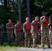 A group of military personnel in camouflage uniforms are standing in a line outdoors, wearing red life vests/personal flotation devices marked with the number '2'. They appear to be participating in a water training exercise or safety briefing near a wooded area. An instructor or cadre member on the right appears to be assisting or inspecting a soldier's life vest fitting.