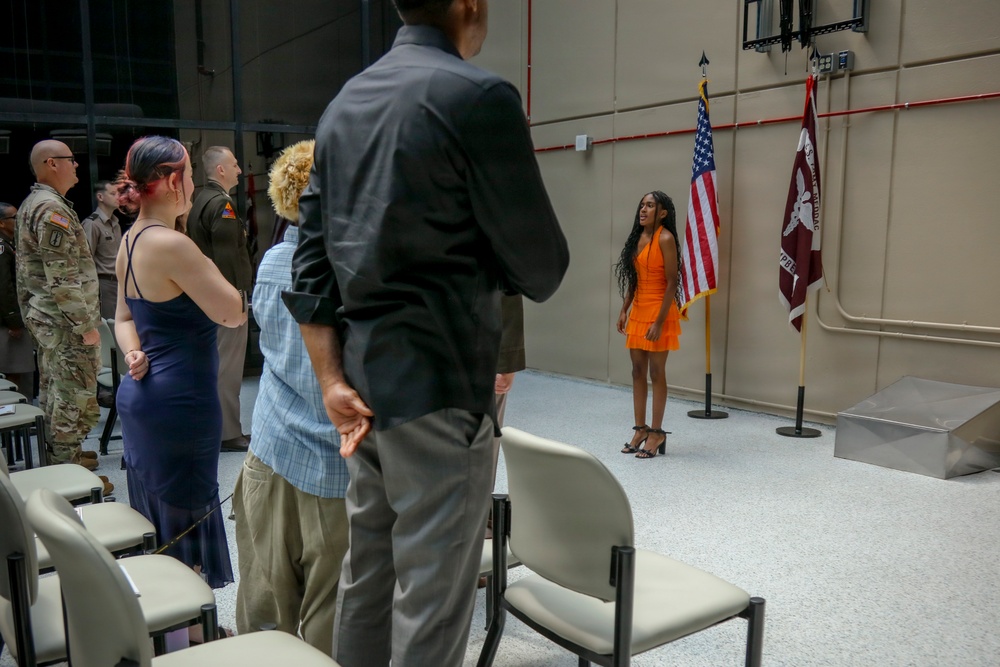 An indoor military ceremony is taking place in what appears to be a garrison or installation hall. Attendees are standing, likely during a formal moment such as the Pledge of Allegiance or National Anthem, with a young woman in an orange dress standing near the American flag and a unit guidon. The setting includes both military personnel in Army Combat Uniforms and civilians in formal and semi-formal attire.