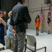 An indoor military ceremony is taking place in what appears to be a garrison or installation hall. Attendees are standing, likely during a formal moment such as the Pledge of Allegiance or National Anthem, with a young woman in an orange dress standing near the American flag and a unit guidon. The setting includes both military personnel in Army Combat Uniforms and civilians in formal and semi-formal attire.