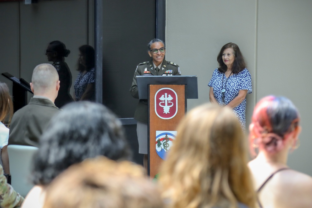 A military officer in olive green uniform is speaking at a podium during a formal ceremony or event. The podium displays an emblem consistent with the U.S. Army Medical Command (MEDCOM), featuring a caduceus symbol on a dark red background. An audience of mixed military and civilian attendees is seated in the foreground.