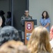 A military officer in olive green uniform is speaking at a podium during a formal ceremony or event. The podium displays an emblem consistent with the U.S. Army Medical Command (MEDCOM), featuring a caduceus symbol on a dark red background. An audience of mixed military and civilian attendees is seated in the foreground.