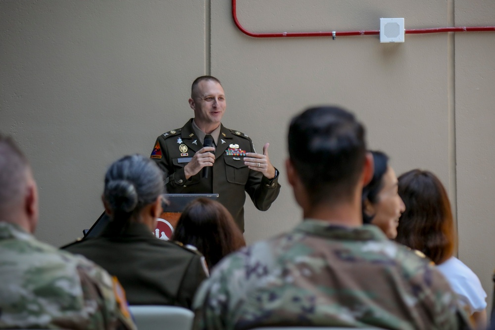 A U.S. Army officer in dress uniform is addressing a seated audience of military personnel and civilians in what appears to be an indoor briefing or ceremony setting. The speaker is standing at a podium and gesturing with his hands while speaking into a microphone. The audience is seated with their backs to the camera, listening attentively.