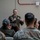 A U.S. Army officer in dress uniform is addressing a seated audience of military personnel and civilians in what appears to be an indoor briefing or ceremony setting. The speaker is standing at a podium and gesturing with his hands while speaking into a microphone. The audience is seated with their backs to the camera, listening attentively.