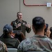 A U.S. Army officer in dress uniform is addressing a seated audience of military personnel and civilians in what appears to be an indoor briefing or ceremony setting. The speaker is standing at a podium and gesturing with his hands while speaking into a microphone. The audience is seated with their backs to the camera, listening attentively.