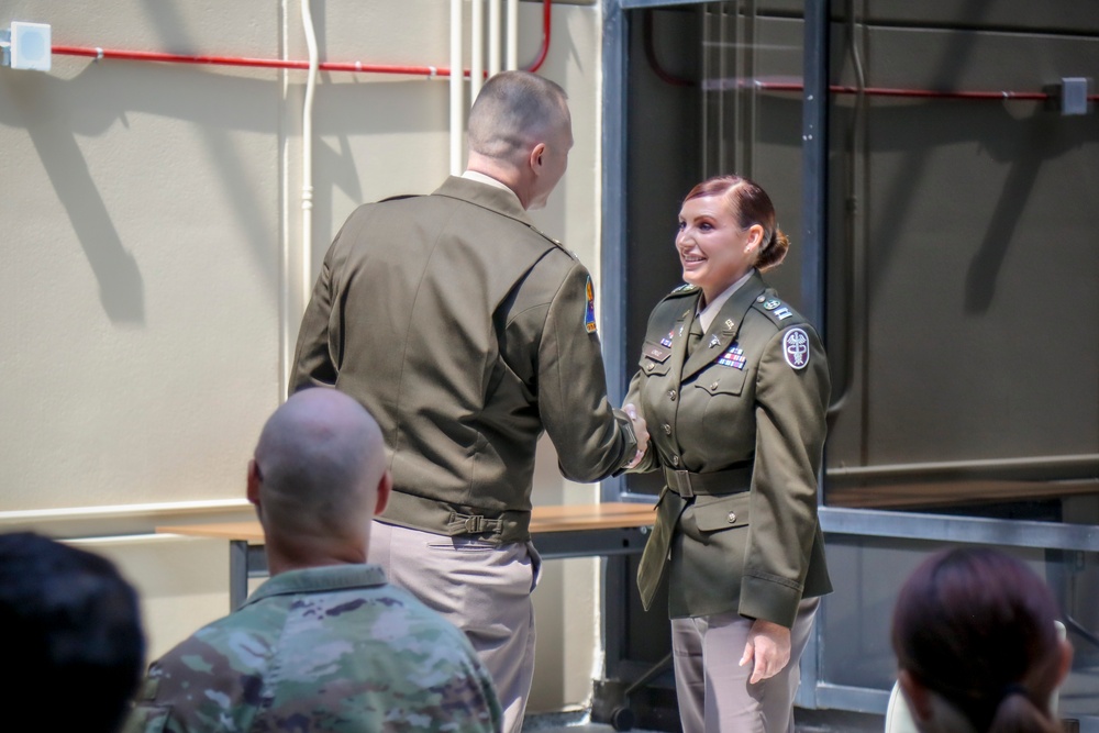 Two U.S. Army soldiers in Army Green Service Uniforms are shaking hands during what appears to be a formal military ceremony. A female soldier facing the camera is smiling while receiving a handshake from a male soldier whose back is to the camera. Several other uniformed personnel are seated in the foreground observing the event.