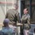 Two U.S. Army soldiers in Army Green Service Uniforms are shaking hands during what appears to be a formal military ceremony. A female soldier facing the camera is smiling while receiving a handshake from a male soldier whose back is to the camera. Several other uniformed personnel are seated in the foreground observing the event.