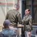 Two U.S. Army soldiers in Army Green Service Uniforms are shaking hands during what appears to be a formal military ceremony. A female soldier facing the camera is smiling while receiving a handshake from a male soldier whose back is to the camera. Several other uniformed personnel are seated in the foreground observing the event.