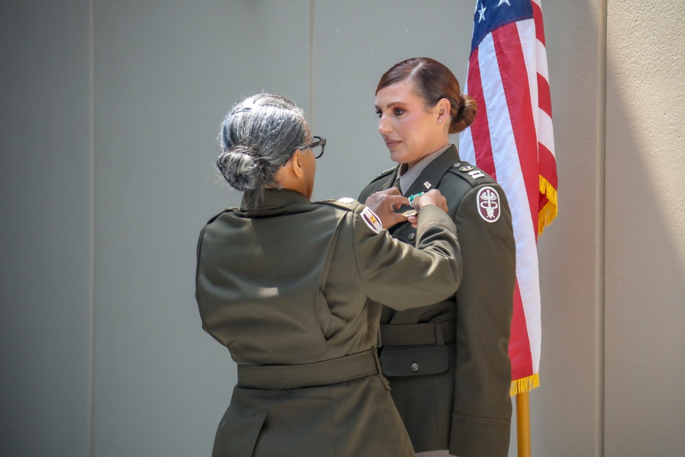 Two individuals in U.S. Army dress green uniforms are participating in a military pinning ceremony. One person, facing away from the camera, is pinning a medal or insignia onto the chest of the other, who faces forward. An American flag with gold fringe is visible in the background.