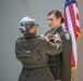 Two individuals in U.S. Army dress green uniforms are participating in a military pinning ceremony. One person, facing away from the camera, is pinning a medal or insignia onto the chest of the other, who faces forward. An American flag with gold fringe is visible in the background.
