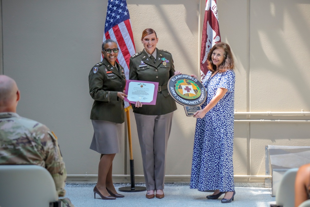 Three individuals pose for a formal recognition ceremony at Blanchfield Army Community Hospital. Two are wearing U.S. Army dress uniforms while a third civilian woman stands alongside them. The group is holding a certificate and a Blanchfield Army Community Hospital commemorative plaque.
