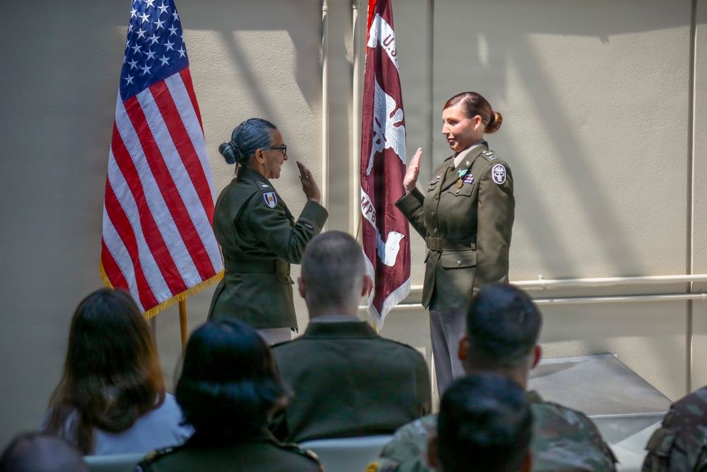 Two female U.S. Army officers are conducting what appears to be a military oath or promotion ceremony, both with right hands raised. They are standing before an American flag and a maroon U.S. Army Medical Corps unit guidon, with an audience of uniformed and civilian personnel seated in the foreground. The setting appears to be an indoor or covered outdoor military facility.