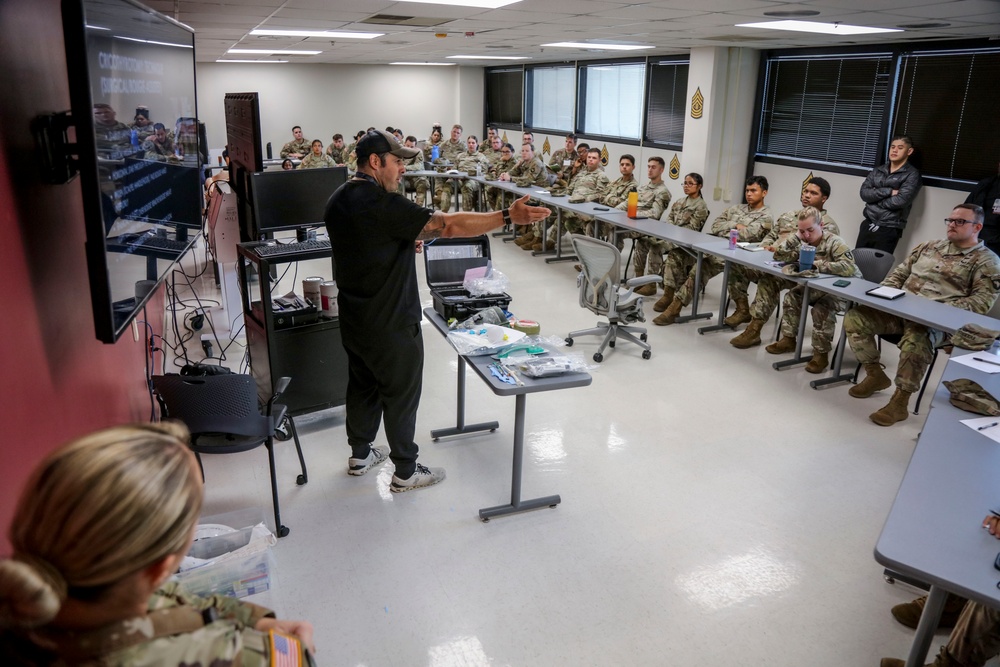 A civilian instructor in black clothing is conducting a training briefing for a large group of military personnel in Army combat uniforms (ACU/OCP pattern). The session appears to be a medical or tactical training class, with medical supplies and equipment visible on a table in the foreground. The soldiers are seated at long tables arranged in a U-shape configuration within a modern classroom facility.