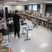 A civilian instructor in black clothing is conducting a training briefing for a large group of military personnel in Army combat uniforms (ACU/OCP pattern). The session appears to be a medical or tactical training class, with medical supplies and equipment visible on a table in the foreground. The soldiers are seated at long tables arranged in a U-shape configuration within a modern classroom facility.