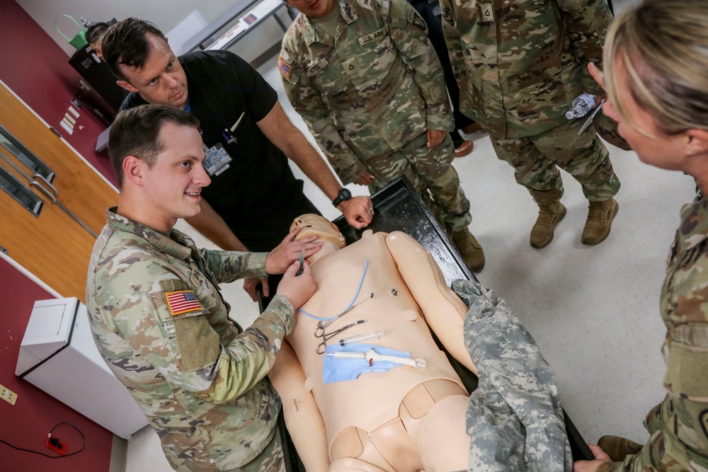 A group of U.S. Army soldiers and a civilian instructor are gathered around a medical training mannequin in what appears to be a simulation lab or medical training facility. A soldier in OCP (Operational Camouflage Pattern) uniform is performing a hands-on medical procedure demonstration on the mannequin while others observe. Various medical instruments are laid out on the mannequin's torso, suggesting a surgical or trauma care training exercise.