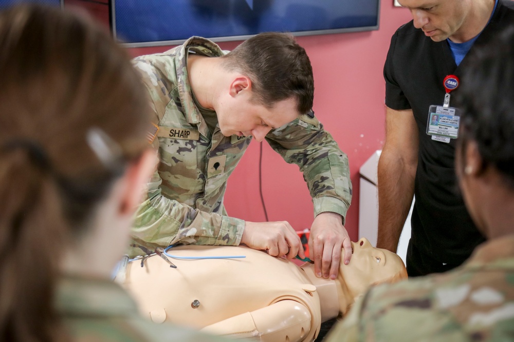 A U.S. Army soldier named 'Sharp' is performing a medical training exercise on a CPR/medical mannequin in what appears to be a simulation training room. The soldier is leaning over the mannequin and appears to be practicing an airway or intubation procedure. A civilian medical instructor wearing scrubs and a badge is observing the training alongside other uniformed personnel.